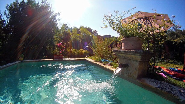 A clear swimming pool with a stone structure on one side, surrounded by greenery and a building with a roof in the background.