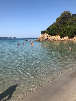 En strand med klart vand, folk der bader, og en klippe med grønt.