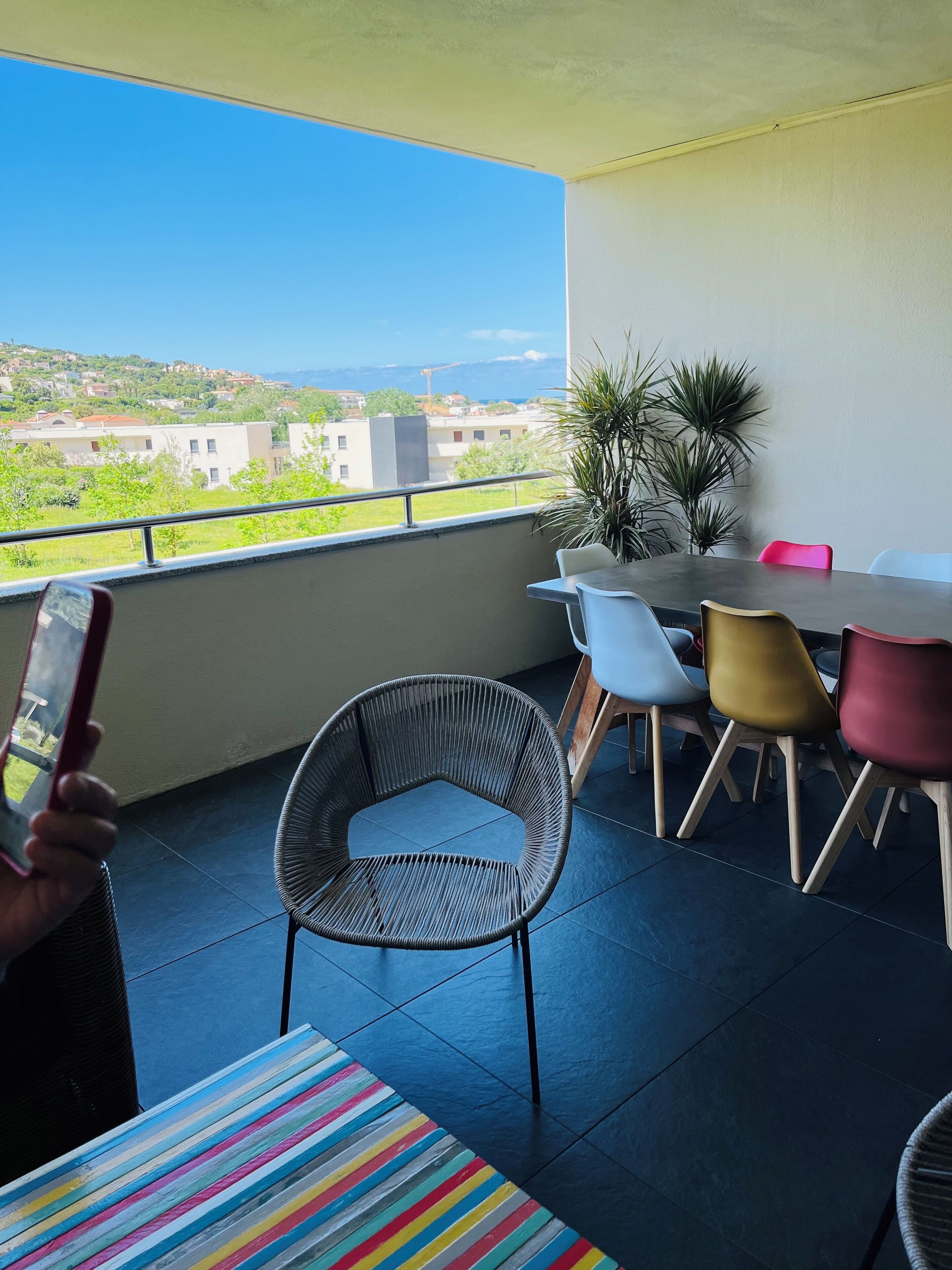 Ein Balkon mit Blick auf Gebäude und einen Baukran, ein Tisch mit Stühlen sowie eine Person, die ein Smartphone hält.