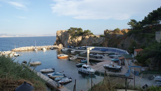 A marina with boats docked, a rocky cliff, and a building with a sign reading "CAPITAÑERIE".