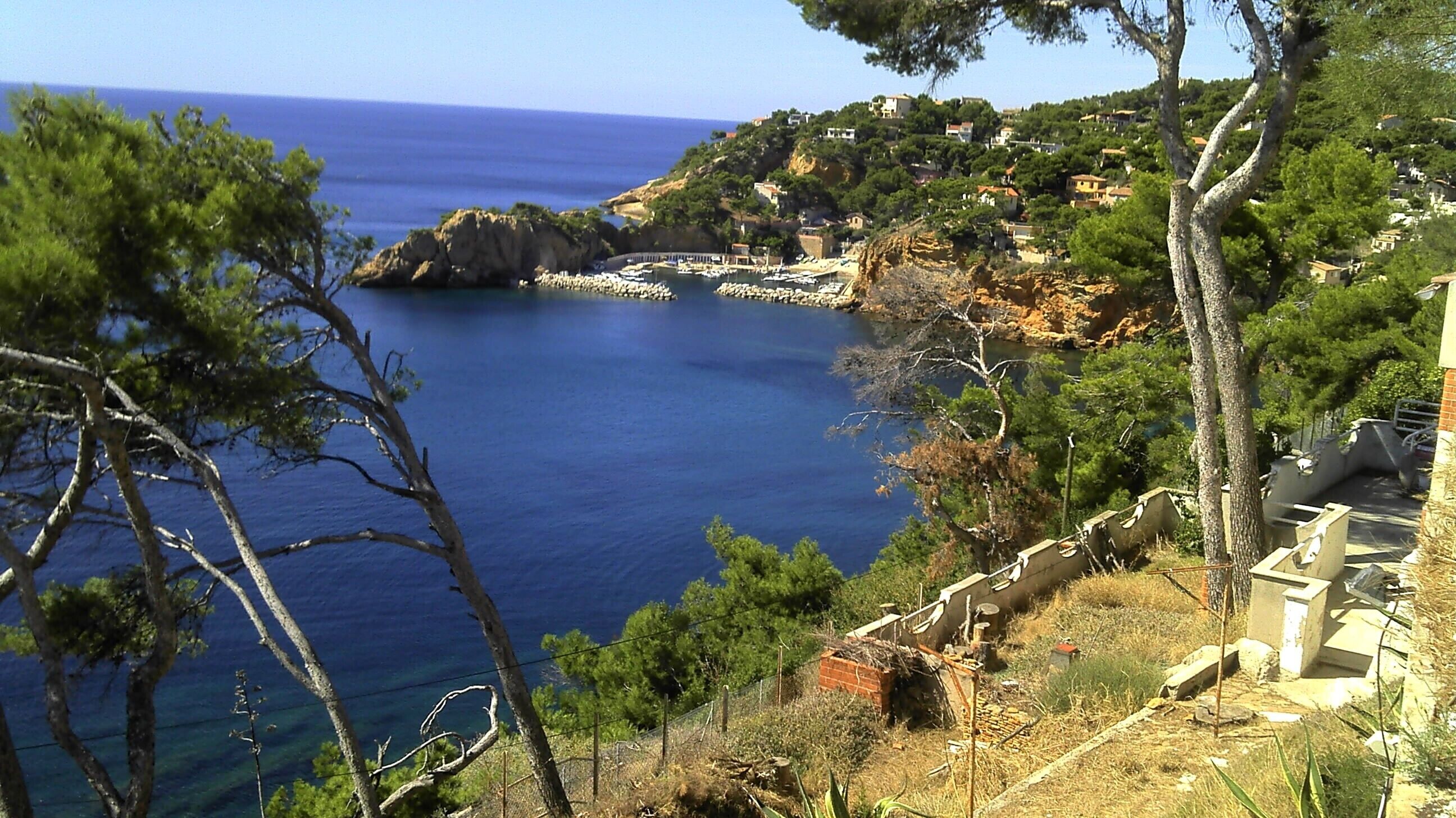 A coastal view with a clear blue sea, rocky cliffs, and a small settlement with buildings.