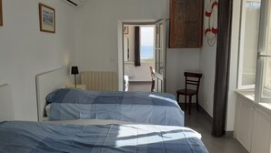 A bedroom with two beds, a chair, a radiator, and a view of the sea through an open door.