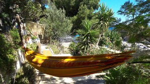 A hammock in a lush garden with a view of a rocky coastline and boats.