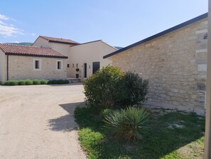 Edifici de pedra amb teulada a dues aigües, carrec de grava i un petit jardí amb un arbust i una planta.