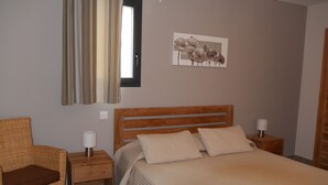 Un dormitorio con una cama de madera, dos mesitas de noche con lámparas, una silla de mimbre, una ventana con cortinas y una obra de arte colgada en la pared.