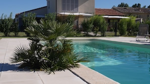 Una piscina con borde de baldosas, rodeada de vegetación y un edificio con techo de tejas.