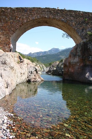 A stone bridge over a clear river with rocky banks and mountains in the background. - Weekly and overnight rentals open all year round (Manso)