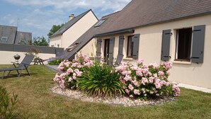 Una casa con techo inclinado, un jardín con flores rosadas y una parrilla de barbacoa.
