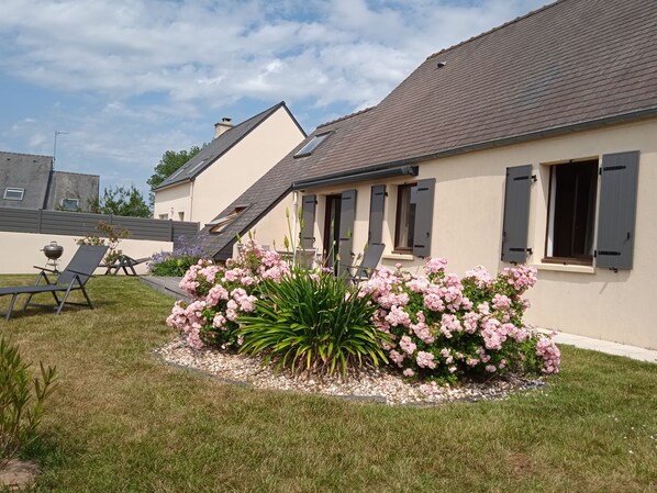 A house with a sloped roof, a garden with pink flowers, and a barbecue grill.