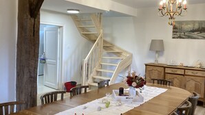 A dining area with a wooden table set for four, surrounded by chairs. A chandelier hangs above, and a painting is on the wall. A wooden staircase is visible in the background. - La Futaie de Chabrehez  (Les Tailles)