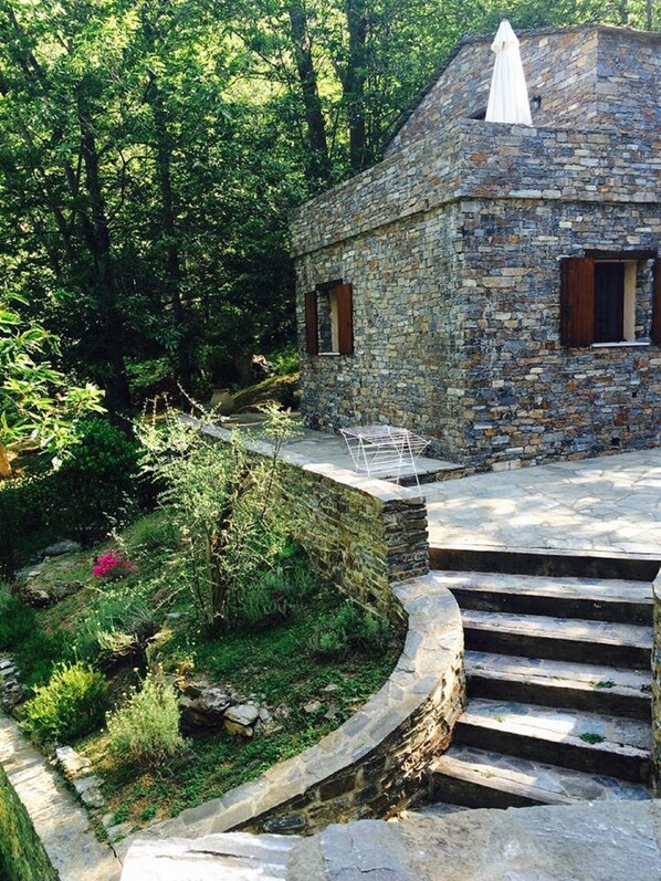 A stone building with a small porch and steps leading up to it, surrounded by lush greenery.