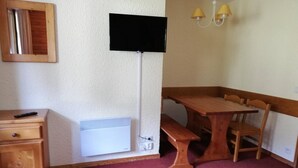 Una habitación con una mesa y banco de madera, un radiador blanco, un televisor montado en la pared y un pequeño mueble de madera.