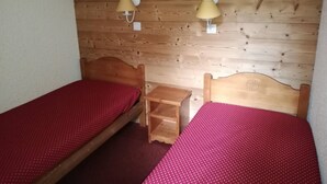 Una habitación pequeña con dos camas individuales, cada una con cabecero de madera y colcha roja. Hay dos lámparas de pared con pantallas beiges.