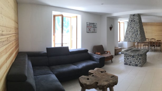 Een woonkamer met een donkere bank, een houten salontafel en een stenen openhaard. Op de achtergrond is een eethoek met een tafel en stoelen te zien.