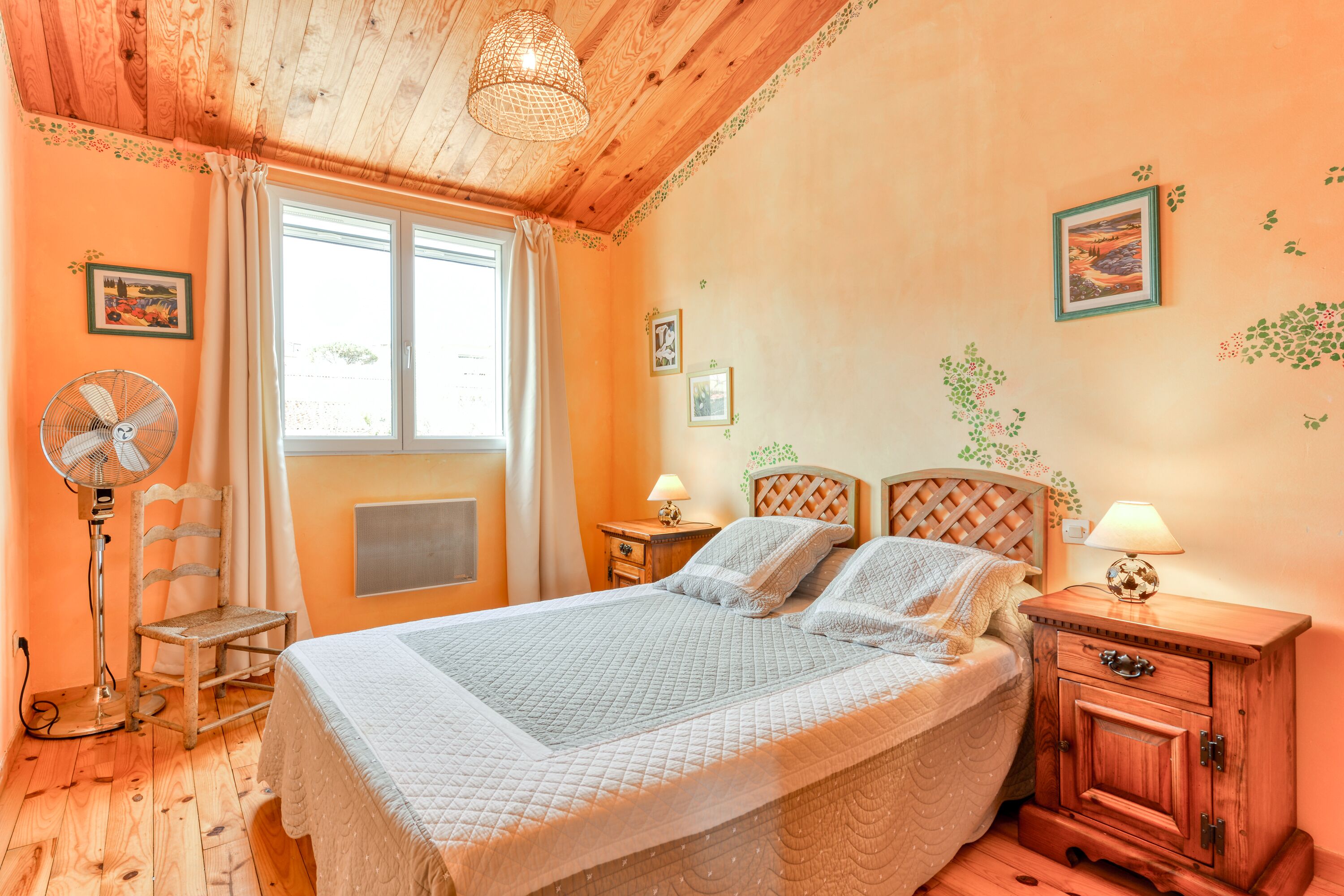 A bedroom with a wooden ceiling, a bed with a quilted bedspread, a nightstand with a lamp, a chair, and a window with curtains.