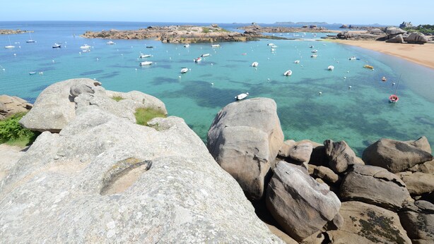 A coastal landscape with clear turquoise waters, large rocks in the foreground, and boats anchored in the bay.