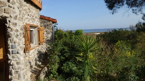 A stone building with wooden shutters, surrounded by greenery and a view of the sea.