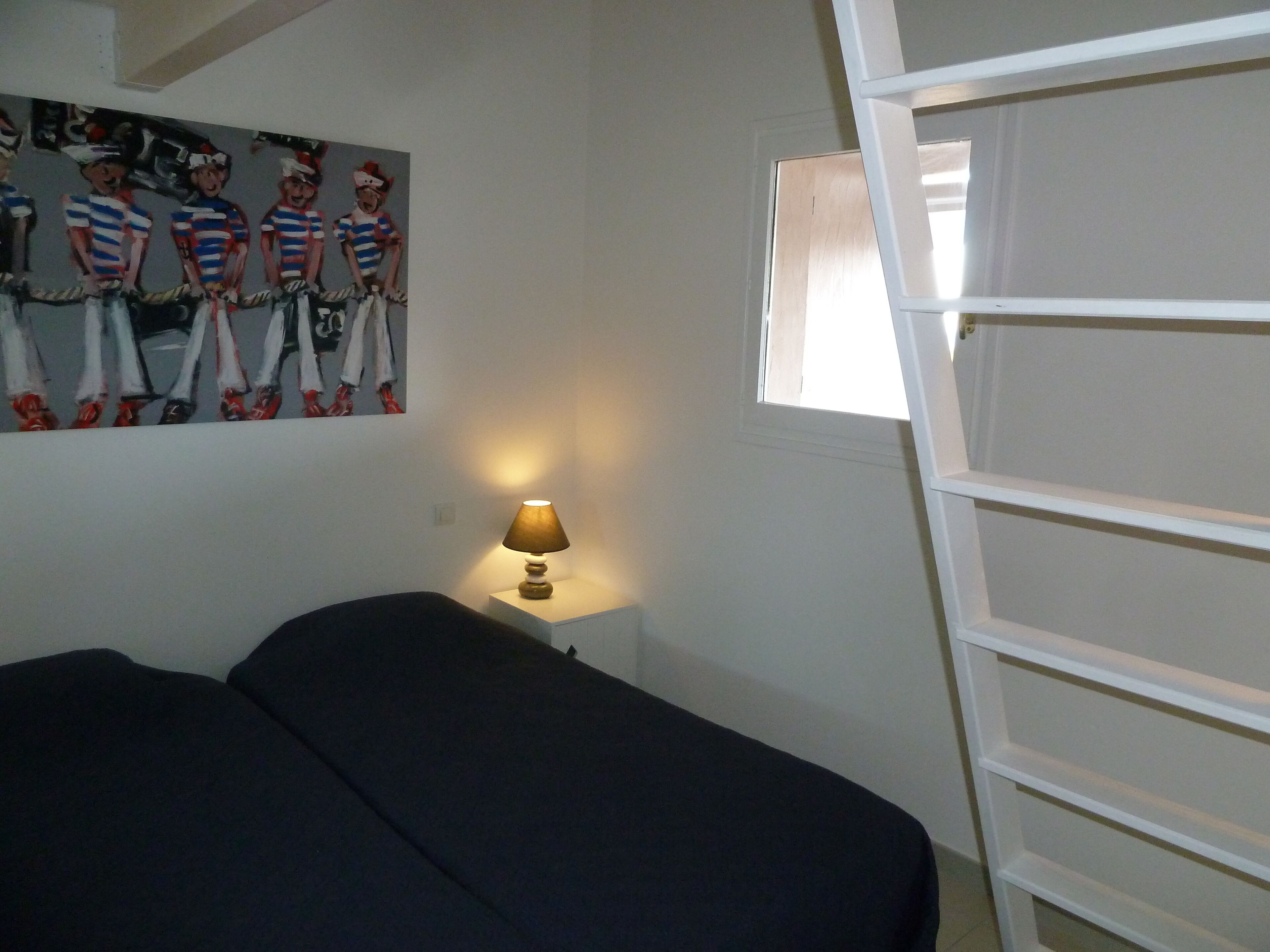 Une chambre à coucher avec un lit, une table de chevet avec une lampe, une fenêtre et un mur orné d’une affiche.