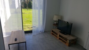Un salón con un televisor sobre un mueble de madera, un control remoto en una mesita y vistas a un área verde a través de una puerta de cristal.
