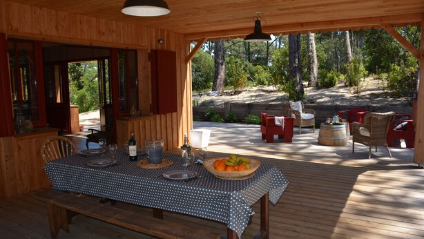 Una terraza de madera con una mesa de comedor preparada para dos personas, rodeada de sillas de mimbre, un banco de madera y vistas a un área boscosa.