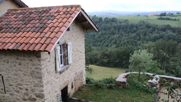 Ein Steinhaus mit rotem Ziegeldach, einem kleinen Fenster und einem Garten mit Steinmauer und Baum.