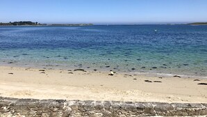 A beach with clear water, a stone wall in the foreground, and distant land with buildings.