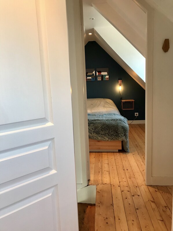 Une chambre avec un plafond en pente, un lit recouvert d’une couverture moelleuse, une lampe fixée au mur et un petit radiateur.