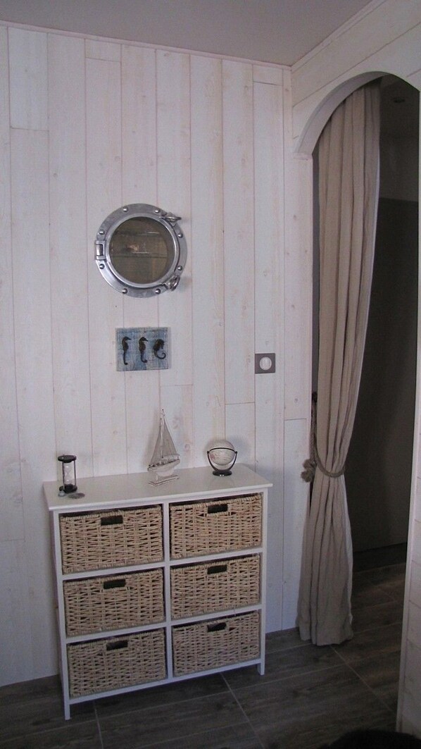 A room with a white wooden wall, a round mirror, a white dresser with wicker baskets, and a beige curtain.