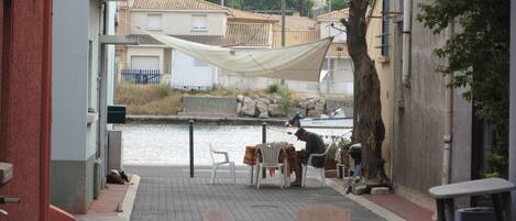 An outdoor seating area with tables and chairs, a body of water in the background, and buildings with tiled roofs.