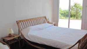 Un lit simple avec un cadre en bois et du linge de lit blanc, dans une chambre comprenant une petite table de chevet, une lampe et une vue sur la verdure par la fenêtre.