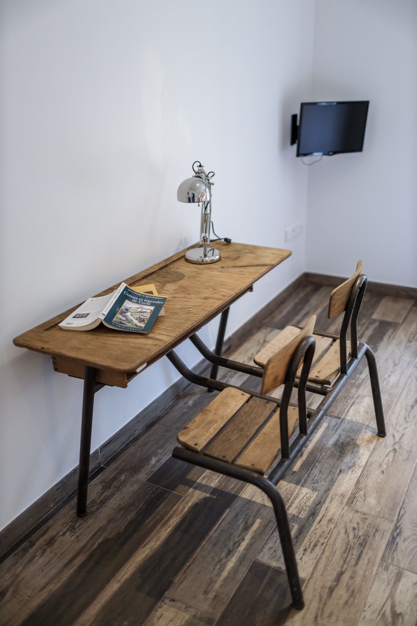 A wooden desk with a lamp, a chair, and a book. A flat-screen TV is mounted on the wall.