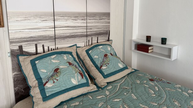 A bedroom with a beach view, a bed with teal and beige bedding, and a white shelf with books and decorative items.