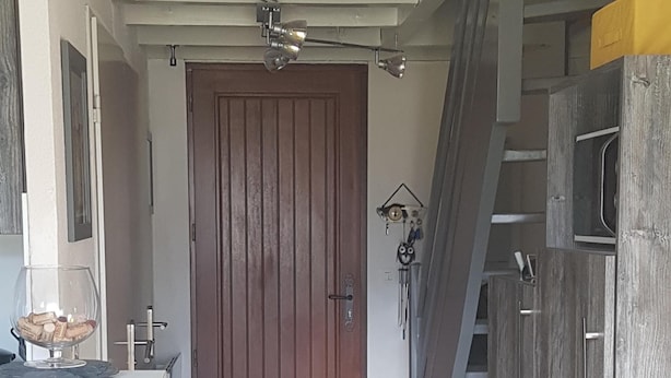 A narrow hallway leading to a wooden door, with a white countertop and a small table with a glass bowl on it. There is a staircase to the right and a wall-mounted light fixture above the door. - A stay by the lake ... (Génos)