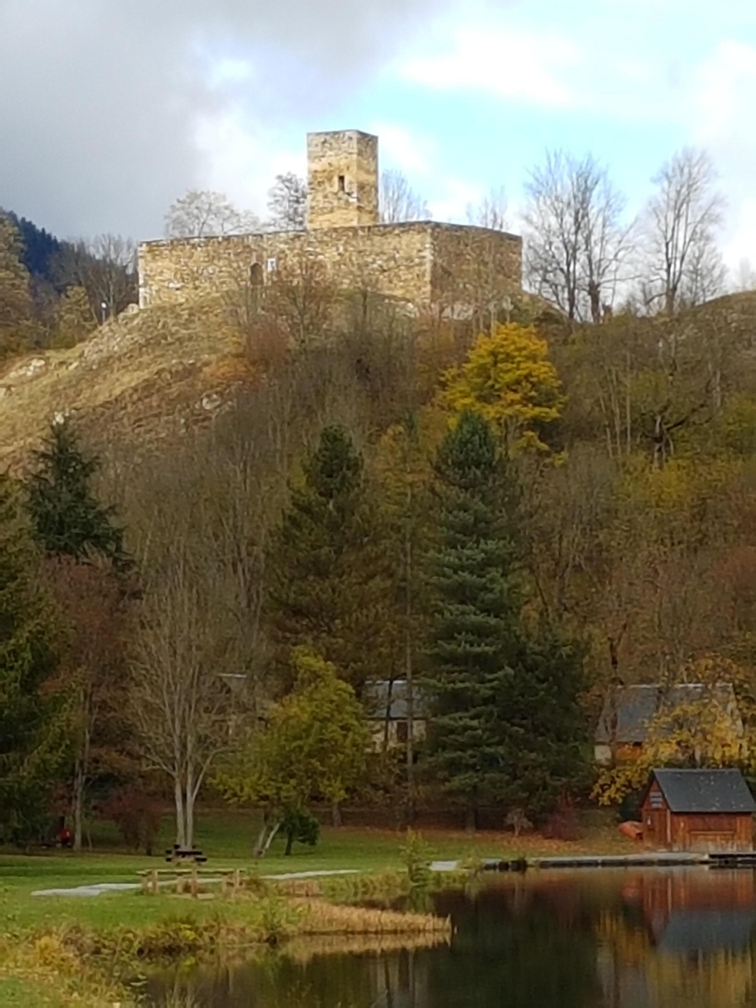 Un château en pierre, perché sur une colline, avec un étang au premier plan et une petite cabane à proximité.