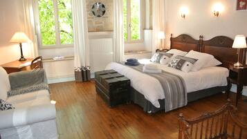 A bedroom with a wooden floor, a bed with white bedding, a bench, a chair, a window with curtains, and wall-mounted lamps.