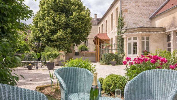 A patio with wicker chairs, a bottle of champagne, and a garden with flowers and a tree.