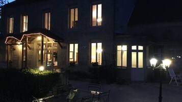 Een stenen gebouw met veel ramen, een overdekt terras met zitplaatsen en een goed verlichte ingang.