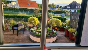 Uitzicht vanuit een raam op een kamer met een houten terras, een stoel en een rond bloembed.