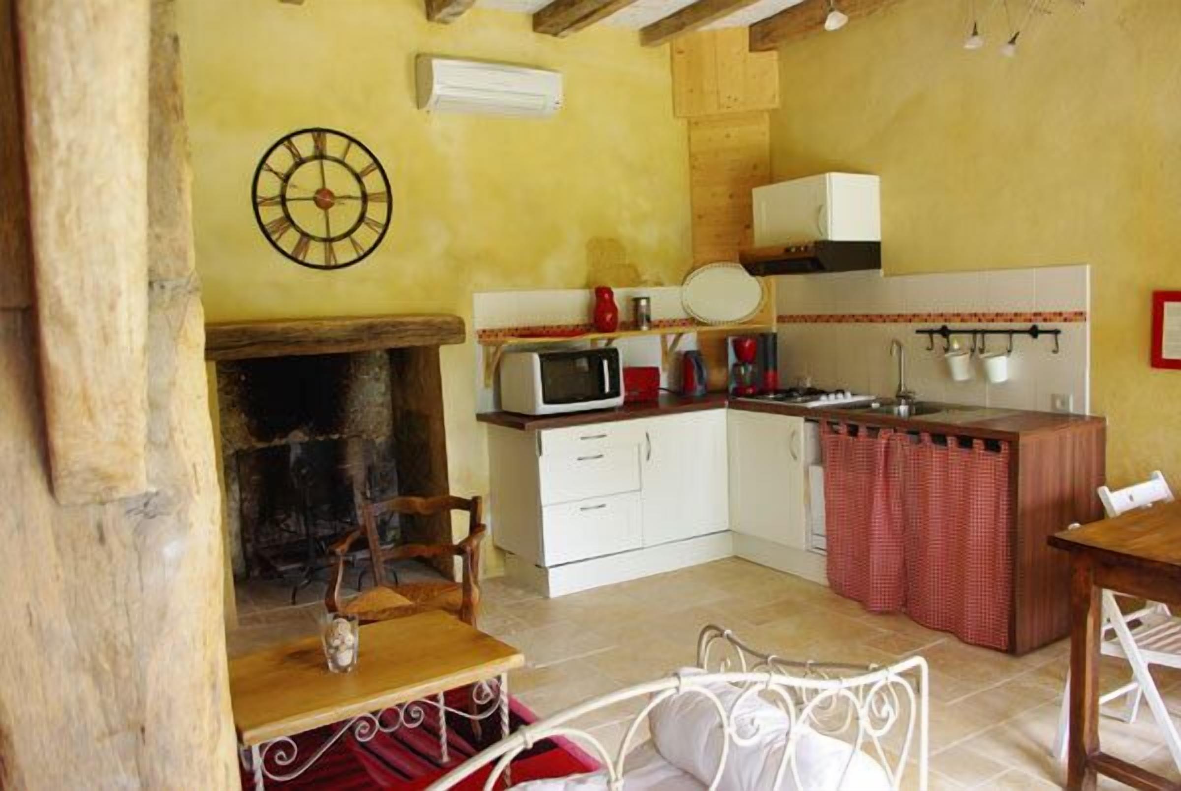 A compact kitchen with a microwave, stove, and sink. There is a wooden table and a chair, and a clock on the wall.