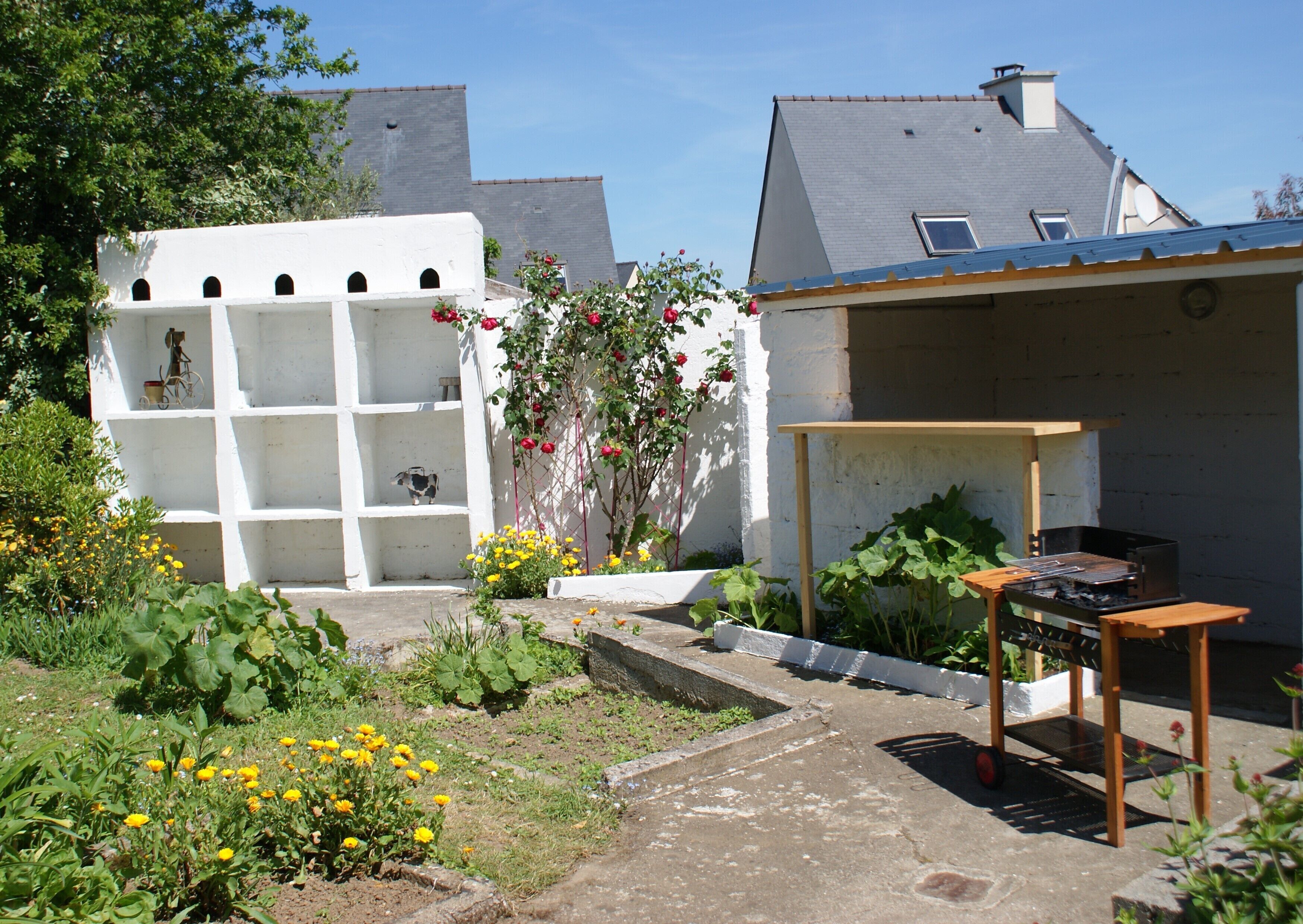 A garden with a barbecue area, a white structure with shelves, and a building with a grey roof.