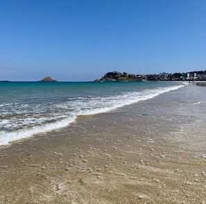 Ein Strand mit klarem Wasser, einem sandigen Ufer und einer entfernten Hügellandschaft mit Gebäuden.