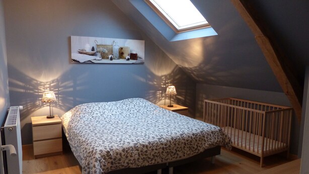 Ein gemütliches Schlafzimmer im Dachgeschoss mit schrägen Wänden, einem Dachfenster, einem Bett mit gemustertem Überwurf, zwei Nachttischlampen, einem Babybett und einem kleinen Regal mit Dekoration.