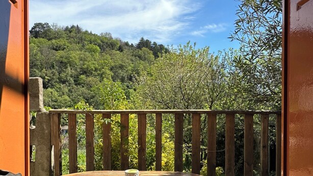 Un balcón abierto con una mesa y sillas de madera, vista a una ladera boscosa y un cielo azul despejado.