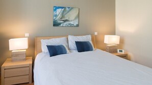 Uma cama bem arrumada com lençóis brancos e duas almofadas, com mesas de cabeceira laterais com abajures e um pequeno relógio. Um quadro de um veleiro está pendurado na parede acima da cama.