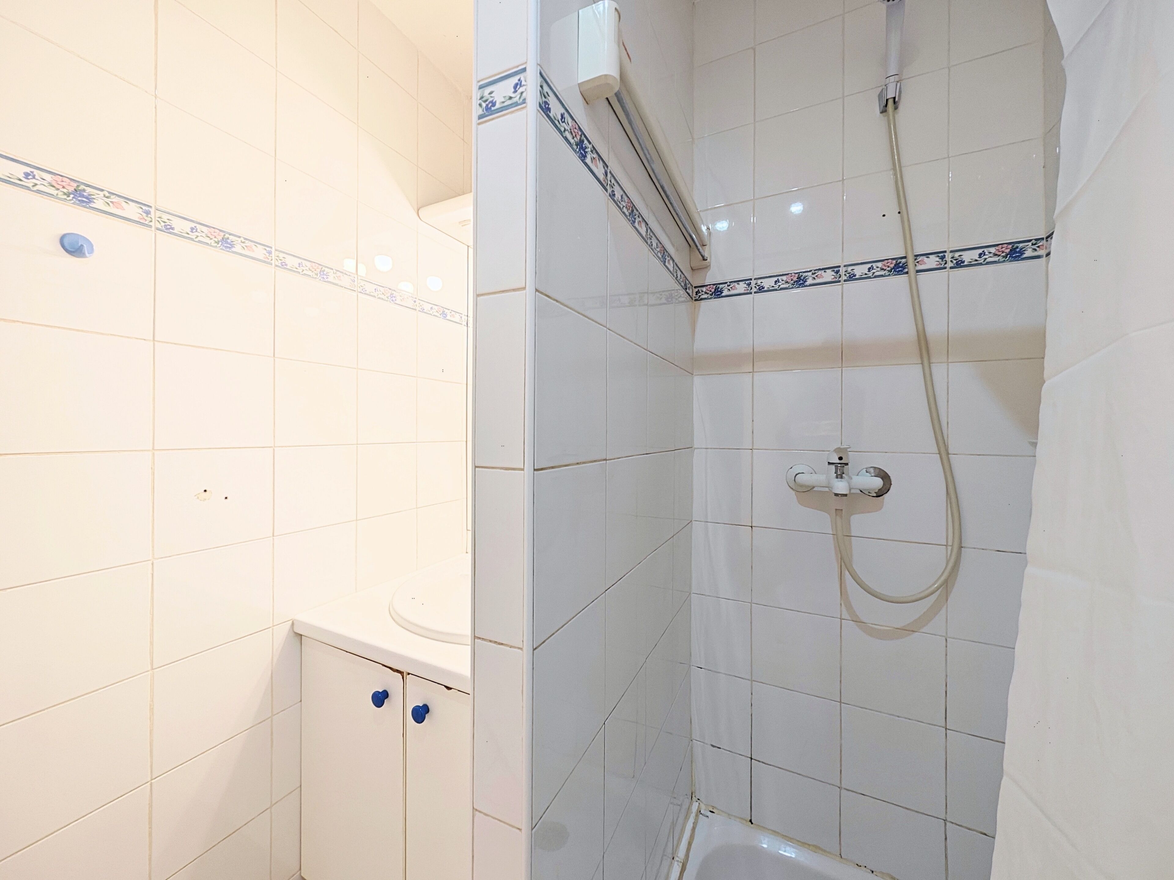 Une salle de bain avec des murs recouverts de carreaux blancs, une douche équipée d’un pommeau de douche à main et une baignoire blanche.
