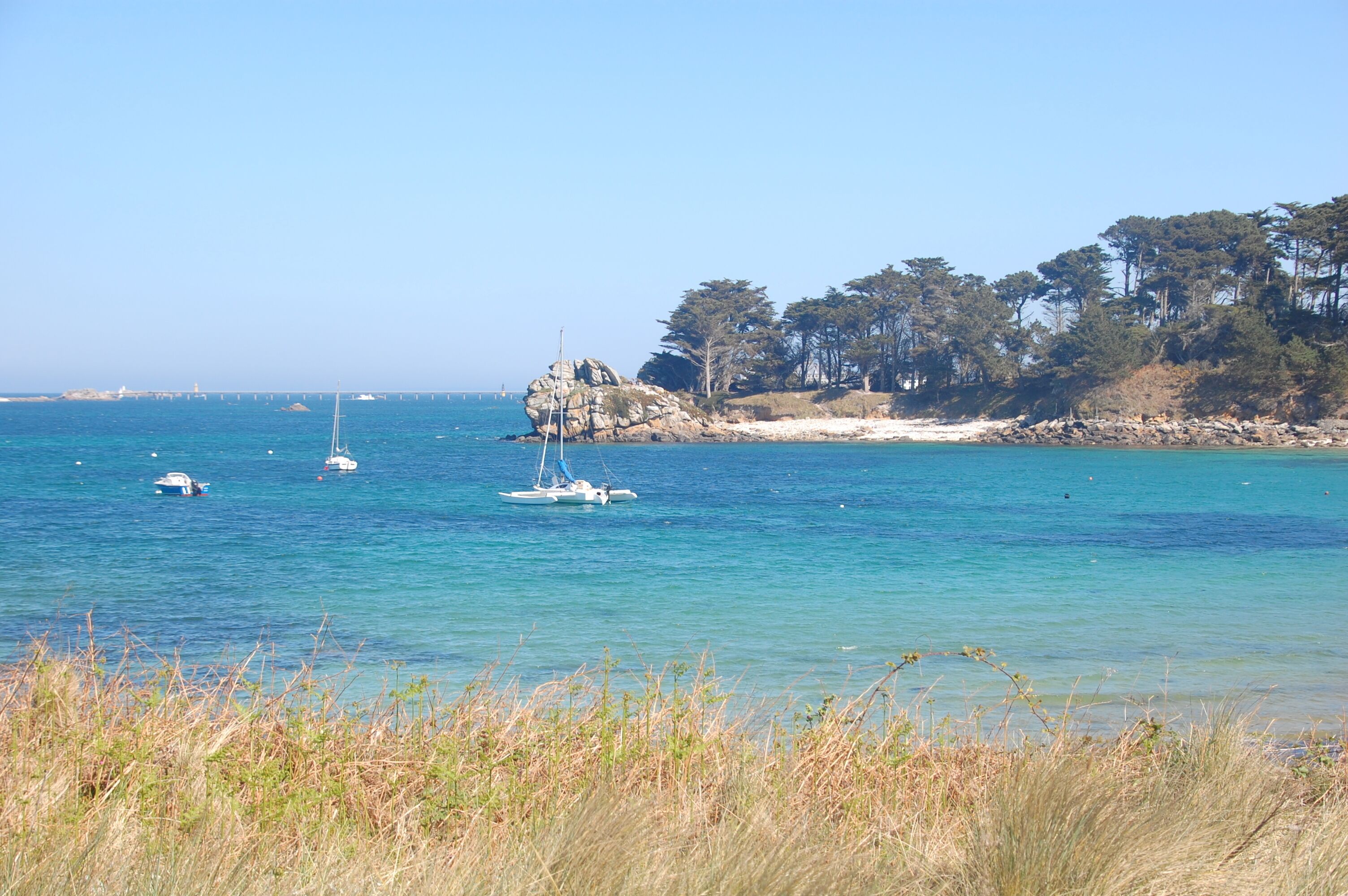 A coastal scene with clear blue waters, boats, and a rocky shoreline with trees.