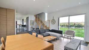 A modern living room with a wooden dining table, grey sofa, and a view of a garden through large sliding doors.
