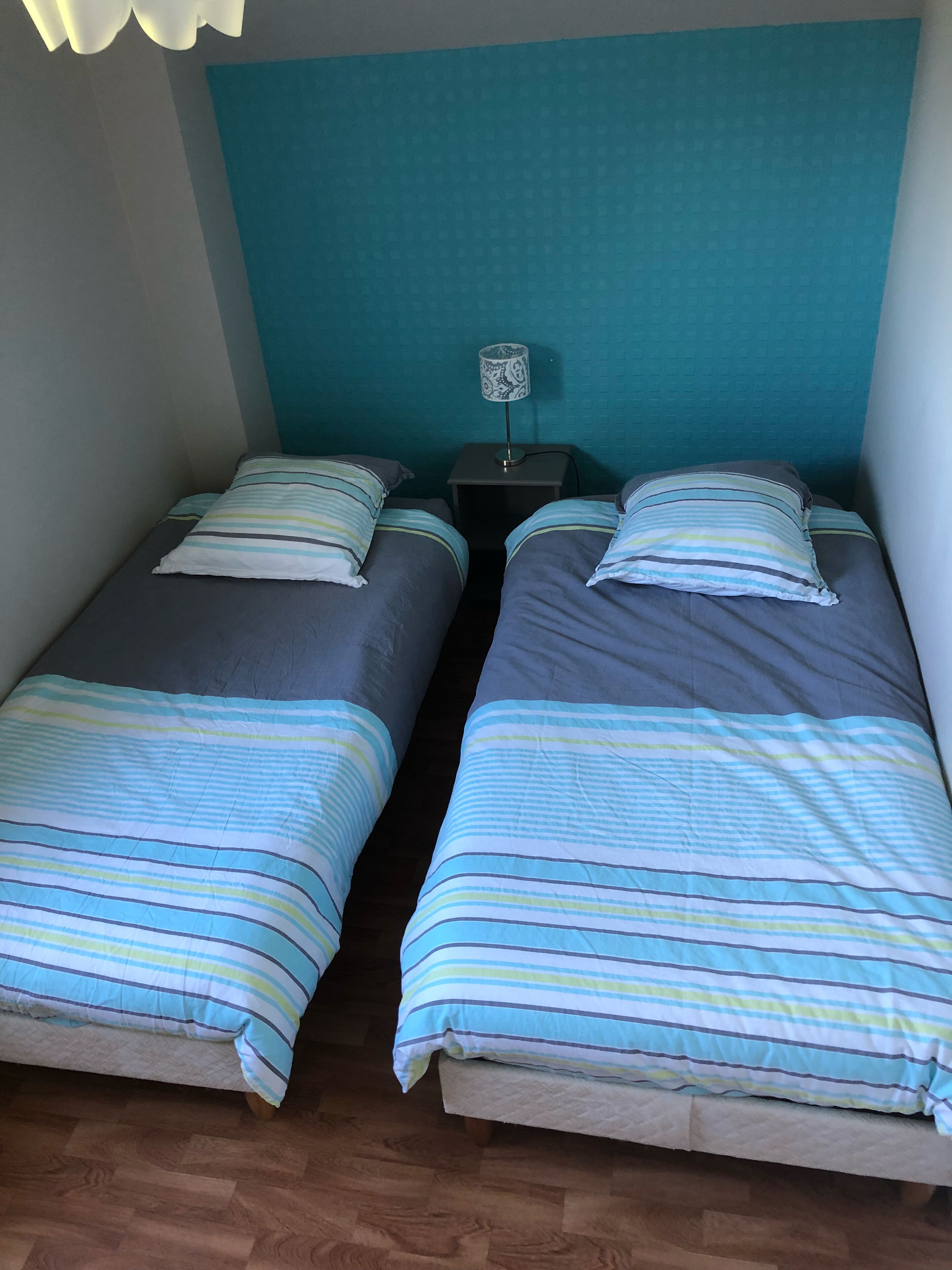 Twee eenpersoonsbedden met gestreept beddengoed, een klein nachtkastje met een lamp, en een muur in een blauwgroene kleur.