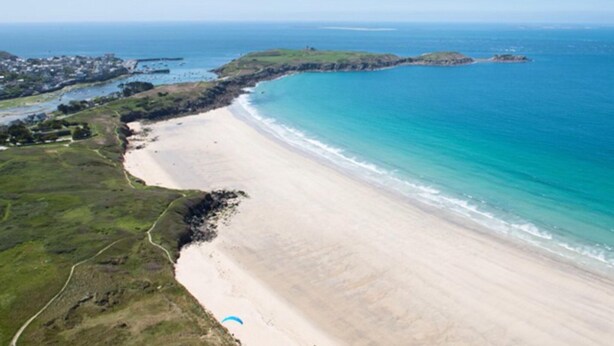 Vista aérea de una zona costera con playa de arena, un pueblo pequeño al fondo y cielo azul despejado.