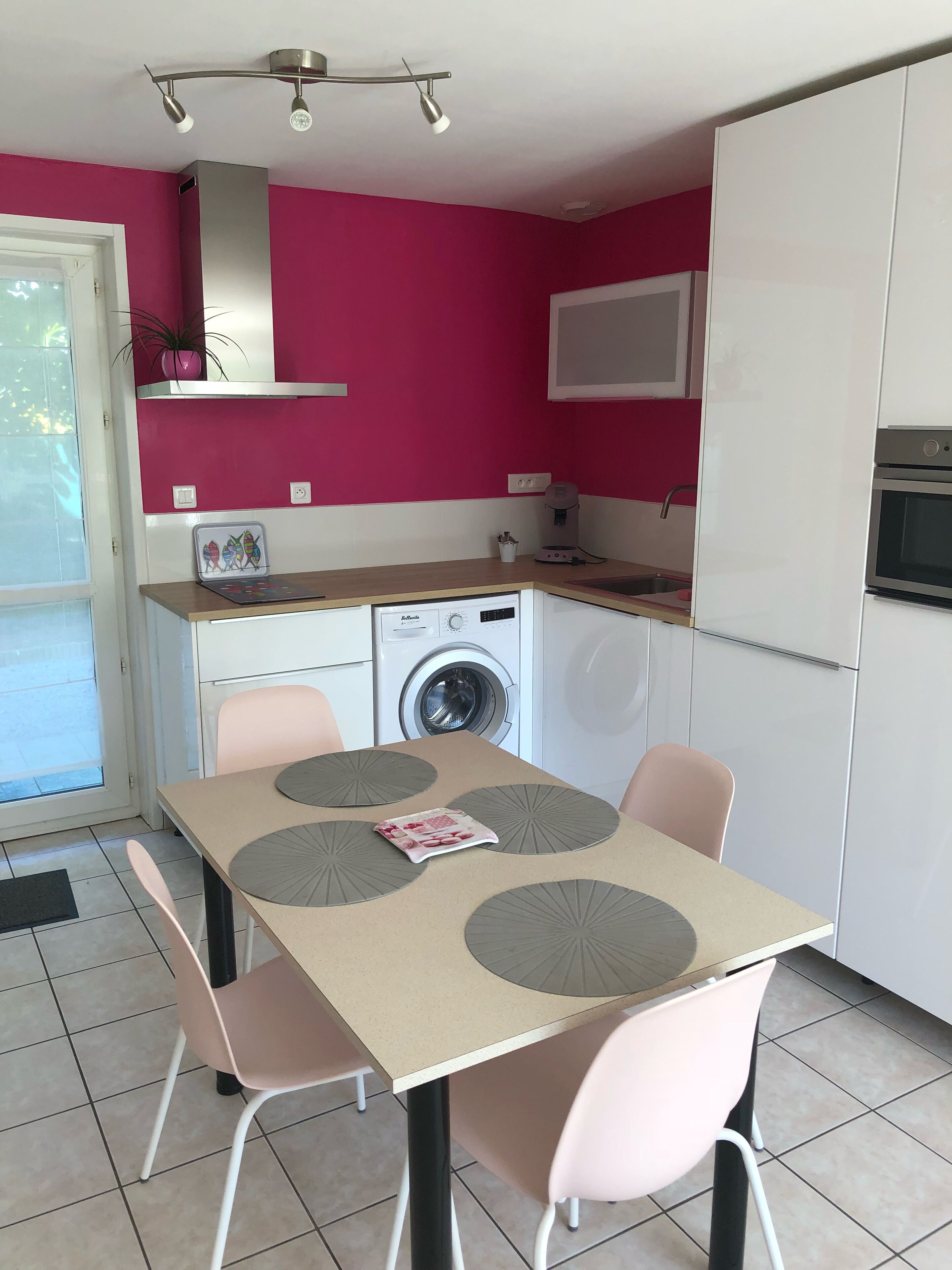 A compact kitchen with a washing machine, a microwave, and a refrigerator. There is a dining table with four chairs and a coffee maker on the counter.